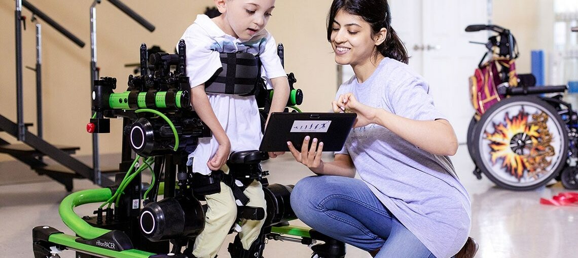Boy with child-sized wearable walking device is guided by researcher.